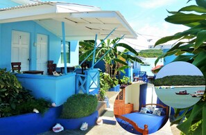 A blue building with numbered doors, a covered outdoor seating area, and a view of boats on the water. - Miller's Guest House (Buccoo)