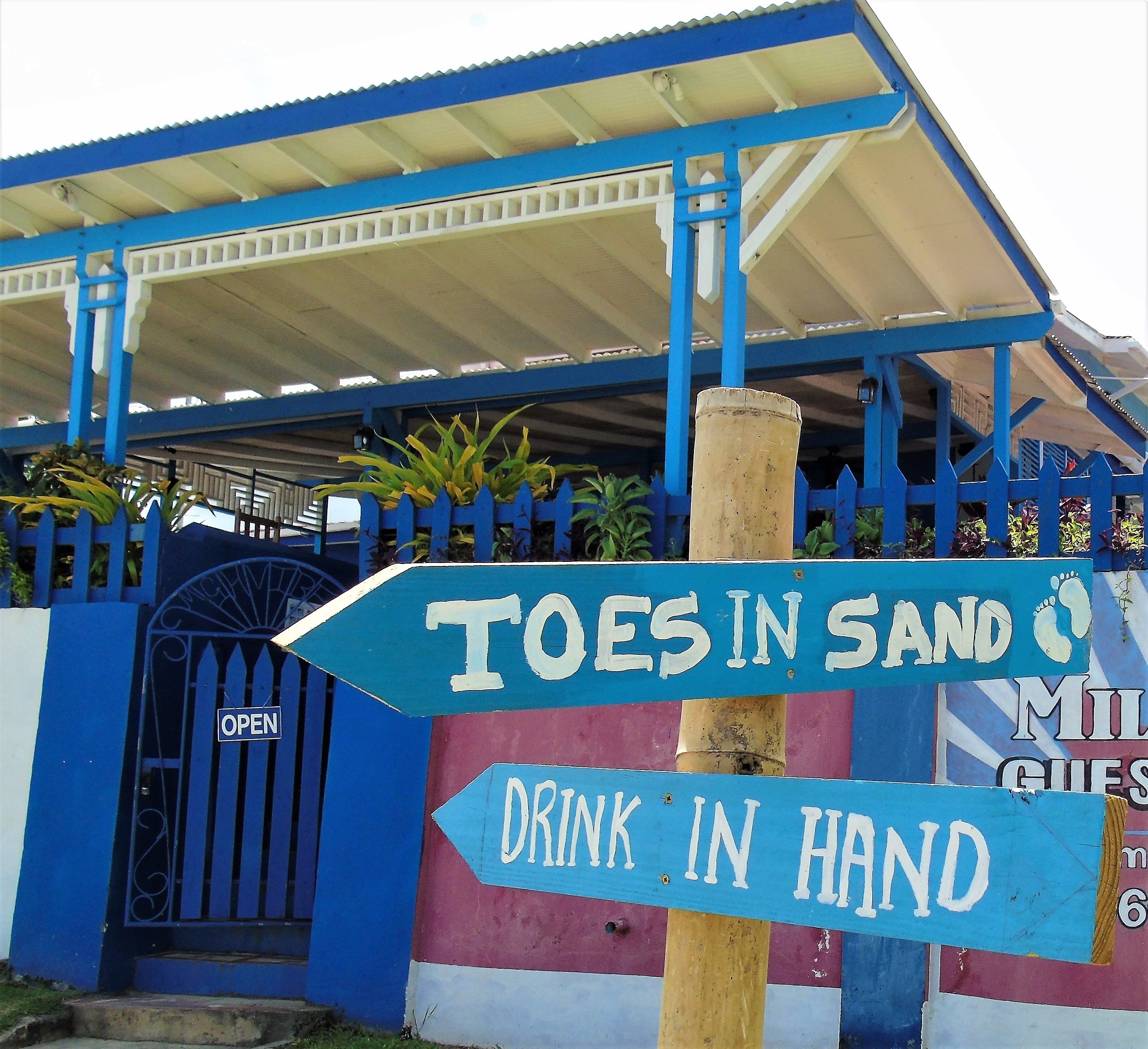 A building with a blue and white facade, a sign that says "TOES IN SAND" and "DRINK IN HAND", and a gate labeled "OPEN".
