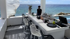 Un espace bar avec vue sur l’océan, comprenant un comptoir où sont exposées différentes bouteilles et différents verres, ainsi que plusieurs chaises blanches.