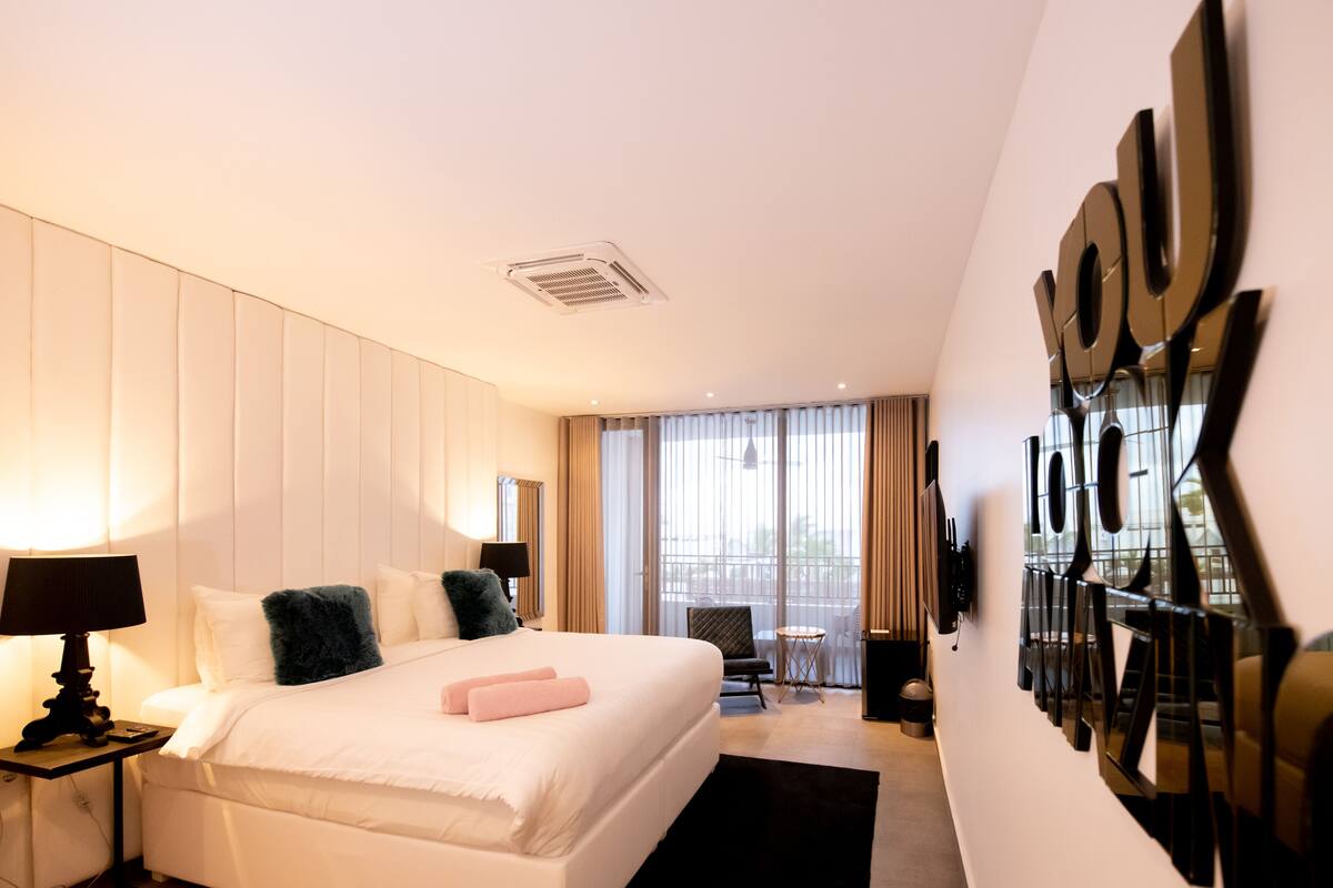 Une chambre d’hôtel avec un grand lit, une table de chevet avec une lampe, une chaise, une petite table et un grand miroir au mur.
