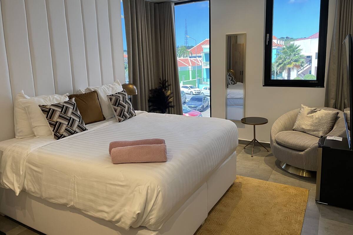 Une chambre d’hôtel moderne dotée d’un grand lit, d’un coin salon avec un fauteuil et offrant une vue sur l’extérieur.