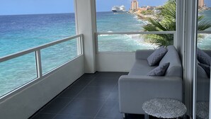 Un balcon avec un canapé blanc et deux petites tables, d’où l’on peut admirer une mer d’un bleu limpide et des bâtiments situés au loin.