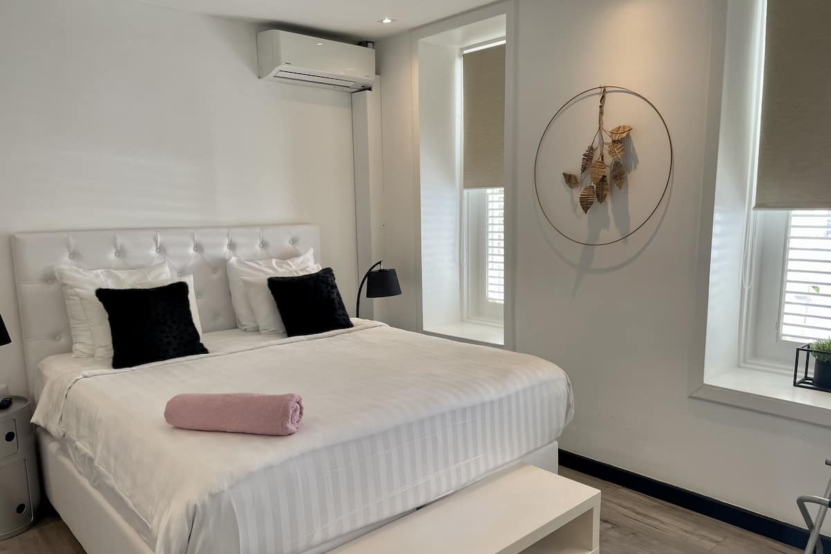 Une chambre moderne avec un grand lit, une table de chevet, un climatiseur fixé au mur, une fenêtre avec des stores et un élément décoratif mural.