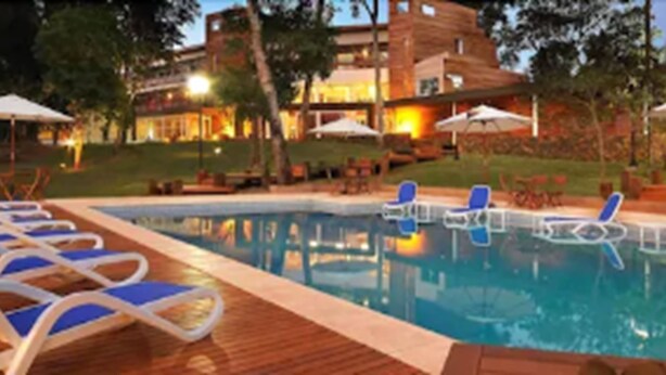 Una piscina con sillones de descanso y sombrillas, rodeada por un edificio de varios pisos y con terraza.