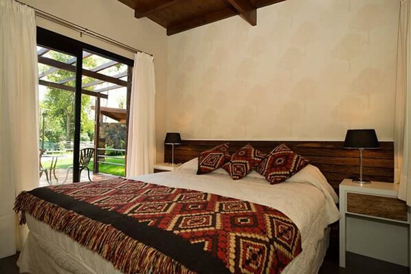 Ein Schlafzimmer mit einem großen Bett, zwei Nachttischen, einem Holzkopfende und Blick auf einen Garten durch eine Glastür.