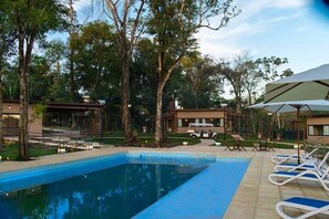 Piscina con sillones de descanso y zona de estar cubierta, rodeada de árboles y edificios.