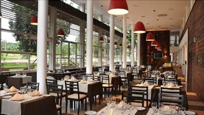 Un restaurante espacioso con techos altos, ventanas amplias e iluminación colgante roja. Las mesas están ordenadas con manteles blancos y preparadas para comer.