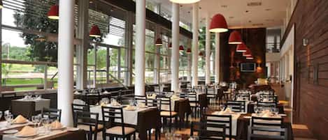Un restaurante espacioso con techos altos, ventanas amplias e iluminación colgante roja. Las mesas están ordenadas con manteles blancos y preparadas para comer.