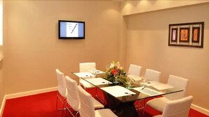 Un salón de conferencias con una mesa de vidrio, sillas blancas, un monitor en la pared y cuadros enmarcados.