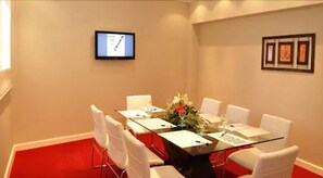Ein Konferenzraum mit Glastisch, weißen Stühlen, einem Monitor an der Wand und gerahmten Kunstwerken.