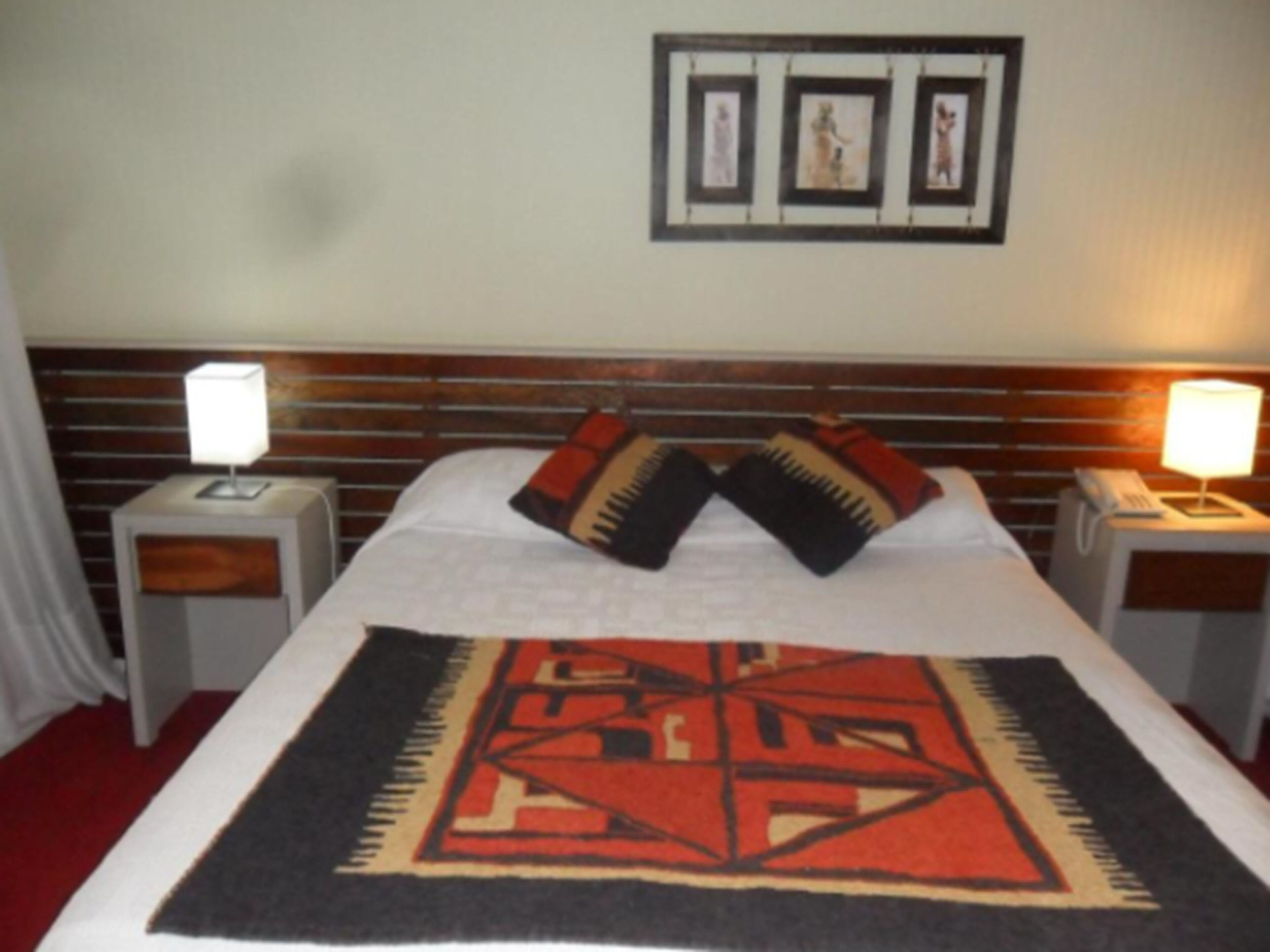 A hotel room with a bed, two bedside tables, a nightstand, a headboard, and a wall with framed pictures.