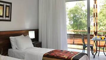 Une chambre d’hôtel avec deux lits, une table de chevet avec une lampe, une grande fenêtre avec des rideaux et un balcon avec une table et des chaises.