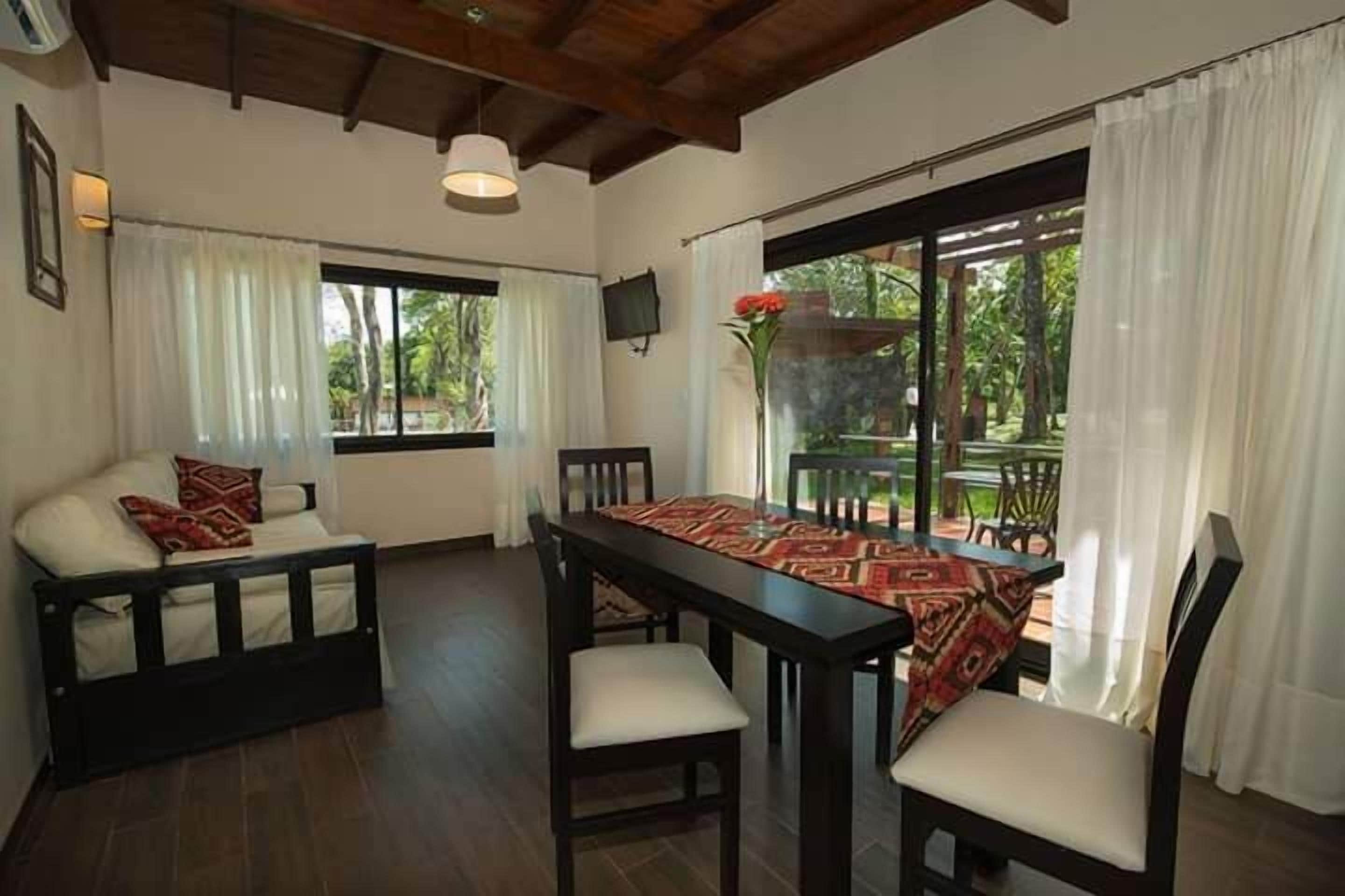 Um quarto com uma mesa de jantar de madeira, cadeiras, um sofá e uma janela grande com cortinas.
