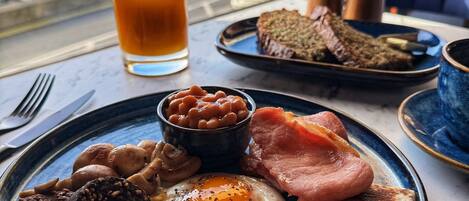 A breakfast plate with sausages, beans, bacon, eggs, and toast, with a glass of orange juice and a view of a cityscape through a window.