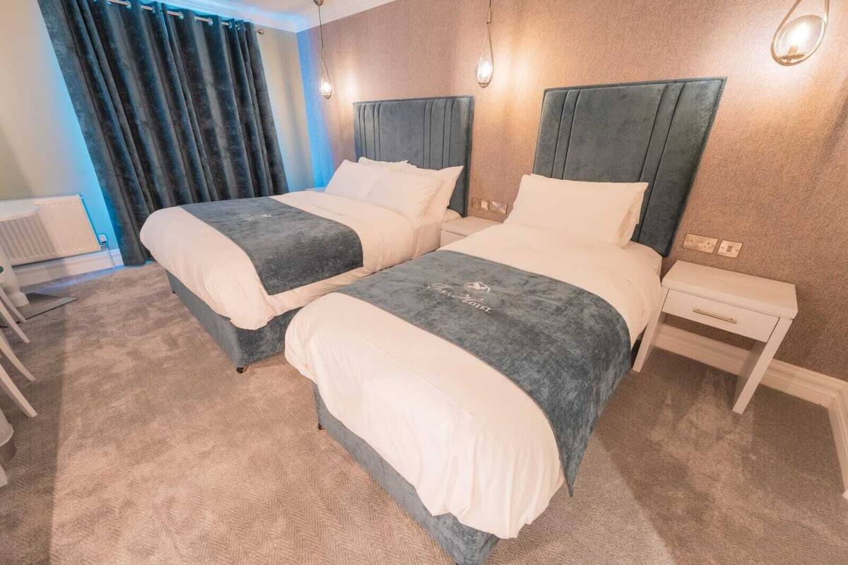 A hotel room with two beds, a small bedside table, a radiator, and a window with curtains.