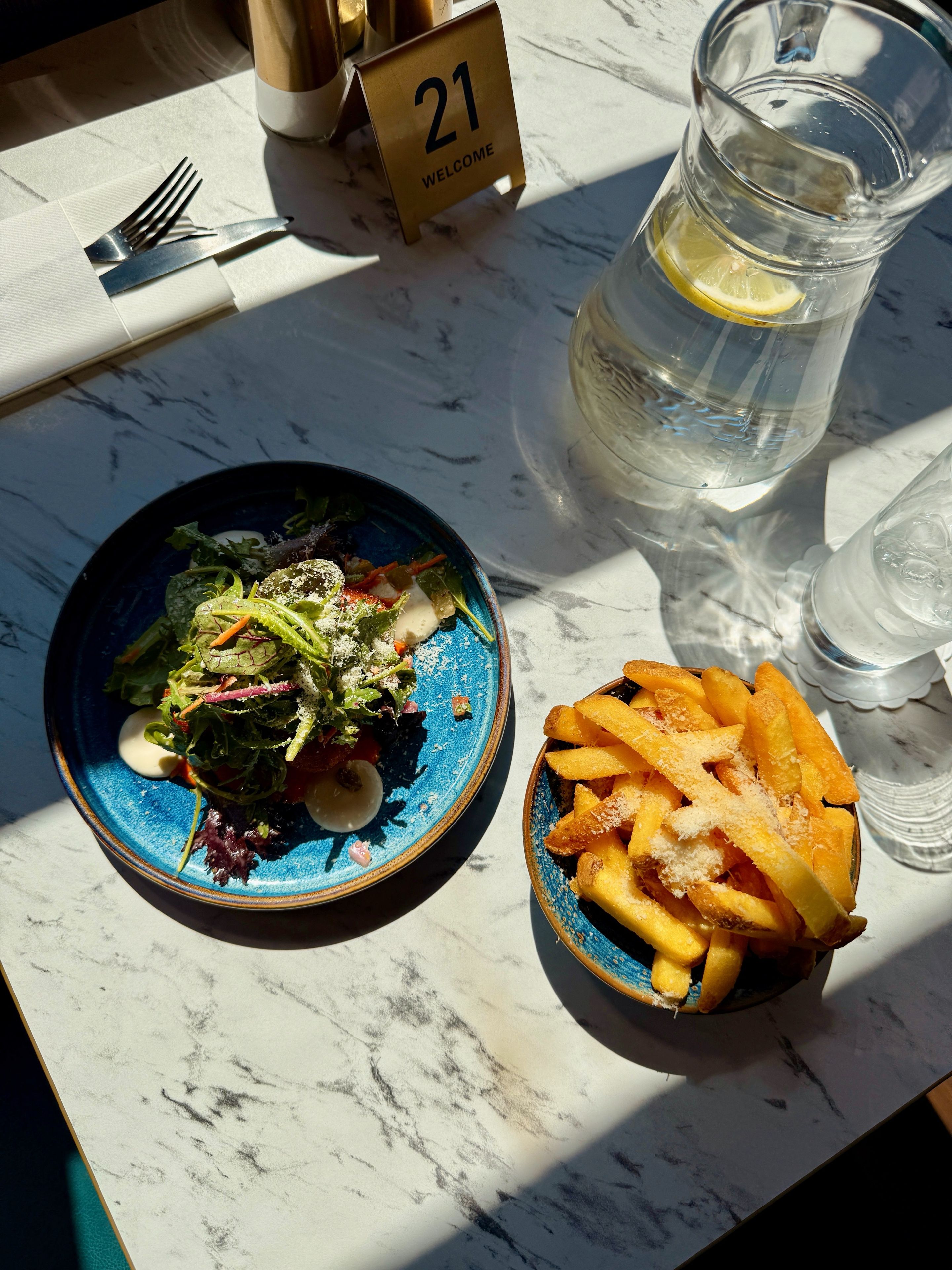 Una mesa con un plato de ensalada azul, un bol de papas fritas, un vaso de agua y una jarra con una rodaja de limón.