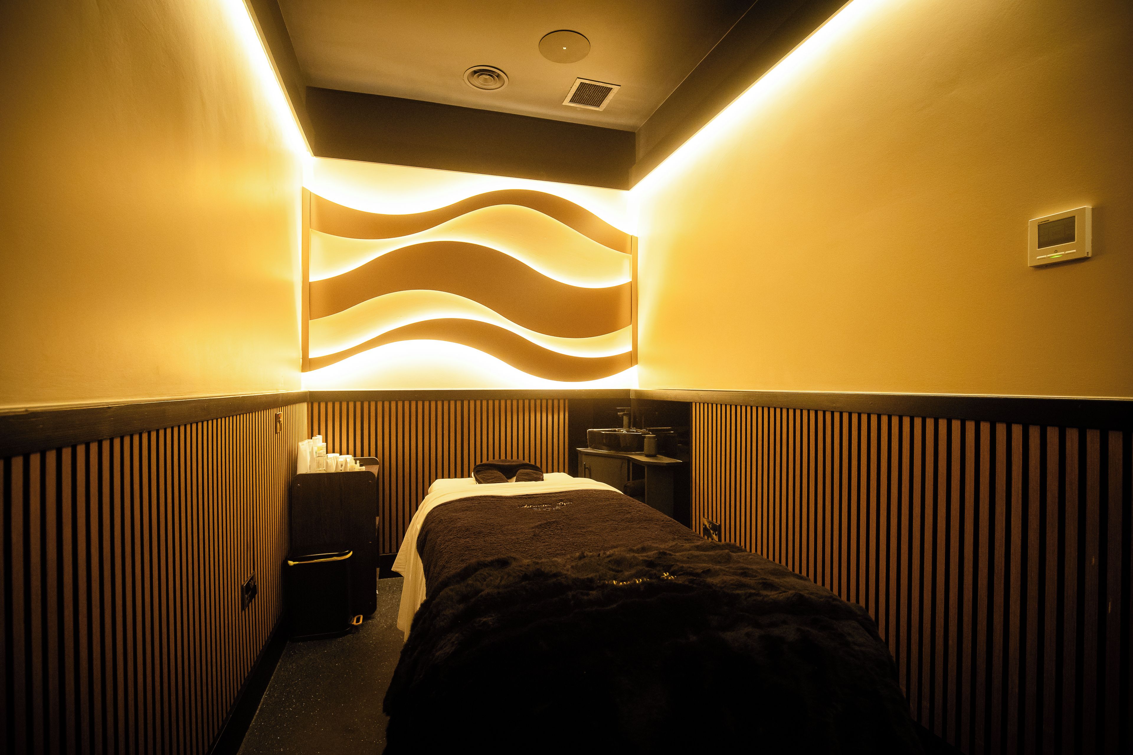 A narrow hotel corridor with a bed at the end, a wall-mounted light fixture, and a thermostat on the wall.