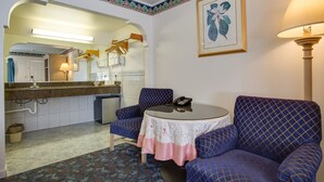A room with a round table, two blue patterned armchairs, a small round table with a lamp, a framed botanical print on the wall, and a mirror above the sink. - Best Whittier Inn (Whittier)