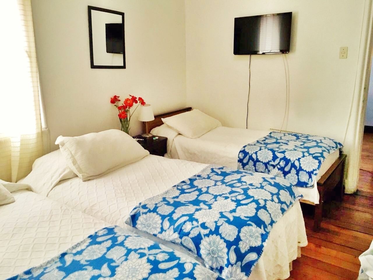 Ein Hotelzimmer mit zwei Betten, einem an der Wand montierten Fernseher und einem Nachttisch mit Lampe und Blumen.
