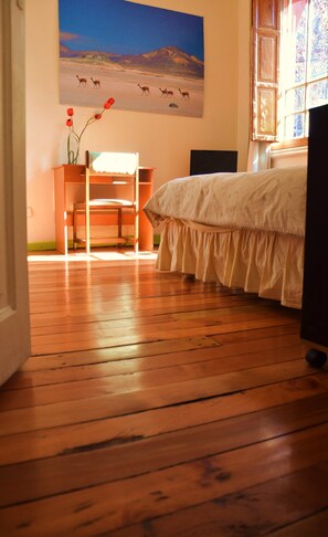 Un dormitorio con piso de madera, una cama con cubrecama blanco, una silla, un escritorio con un jarrón de flores rojas y un cuadro de animales en la pared.