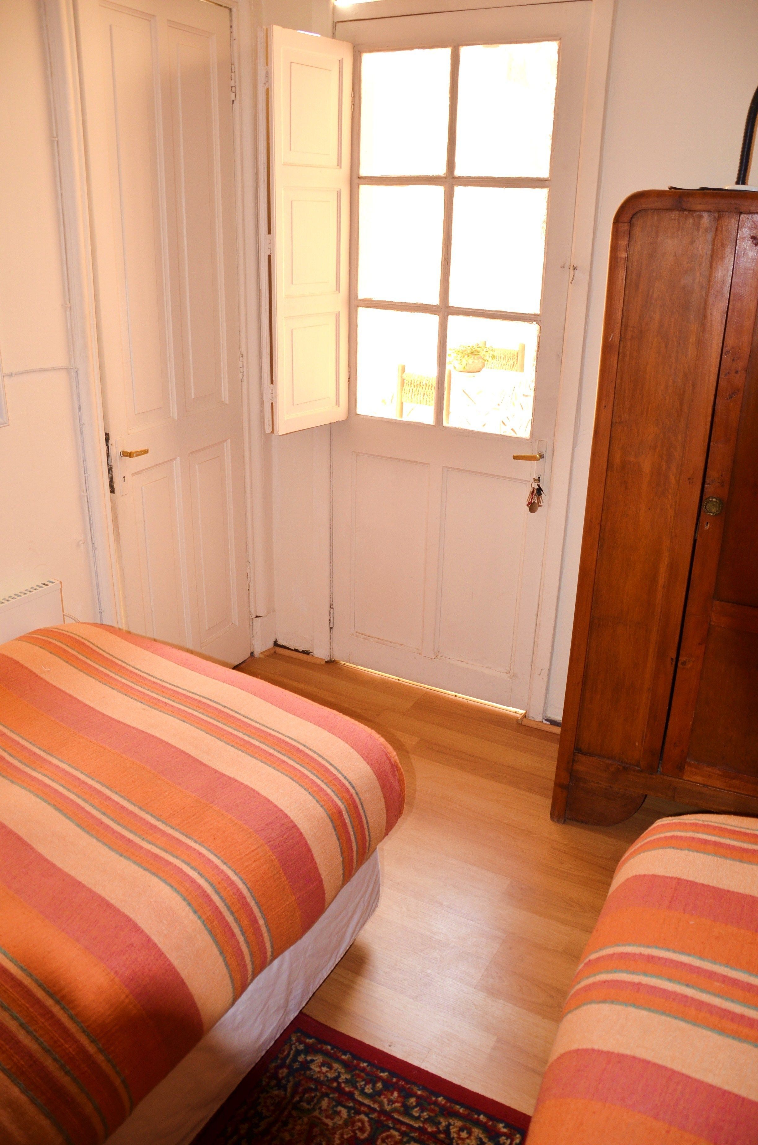 A room with two beds, a wooden wardrobe, and a door leading to a bright area.