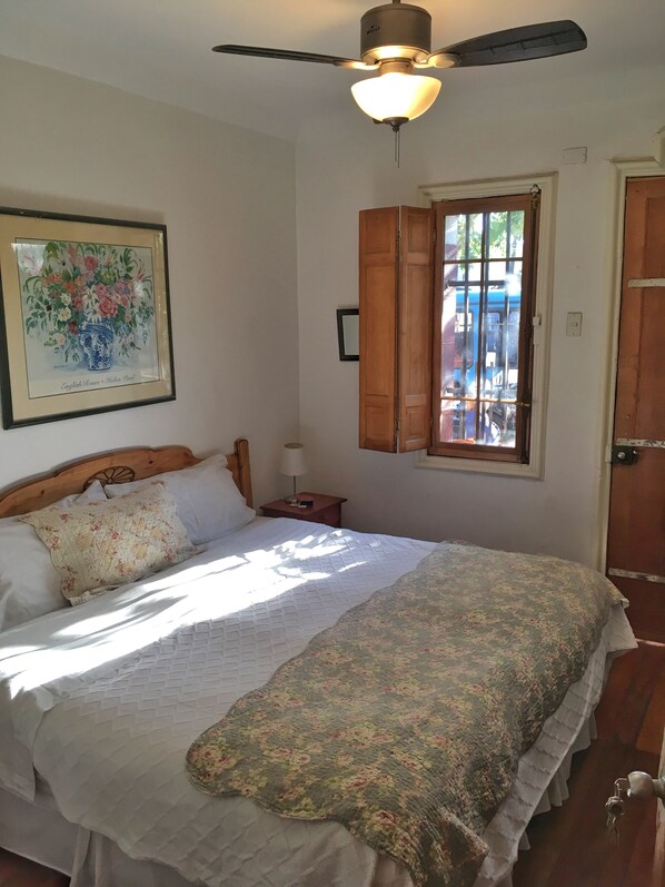 Un dormitorio con cama, ventilador de techo, ventana con contraventanas de madera y una obra de arte floral enmarcada en la pared.