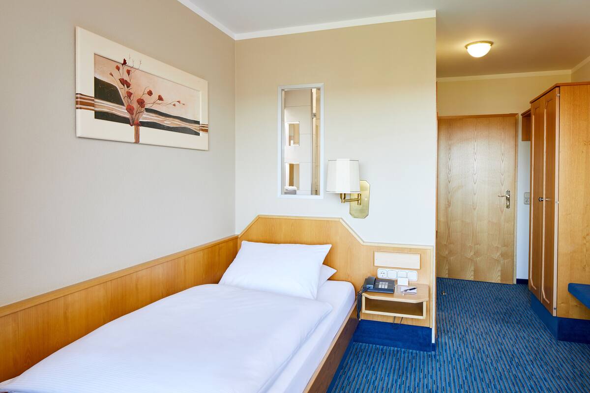 Une chambre d’hôtel avec un lit en bois, une table de chevet avec une lampe, un miroir, une armoire en bois et une œuvre d’art encadrée au mur.