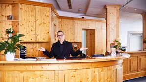 A wooden reception desk with a person standing behind it, a potted plant, and a counter with various items. - Hotel Sonnenhof Dietzenbach (Dietzenbach)