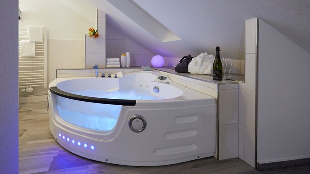 A modern bathroom with a built-in jacuzzi, a towel rack, a bottle of champagne, and a glass of wine on the counter. - Hotel Sonnenhof Dietzenbach (Dietzenbach)