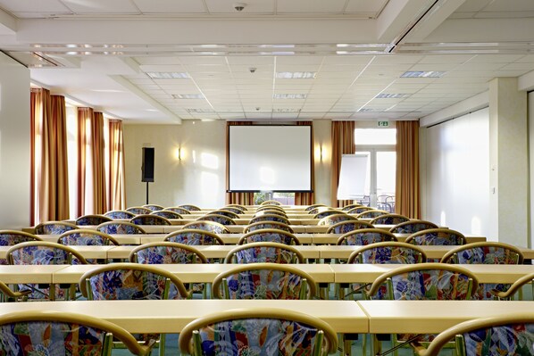 Un salón de conferencias con mesas y sillas dispuestas en filas, una pantalla de proyector y cortinas.