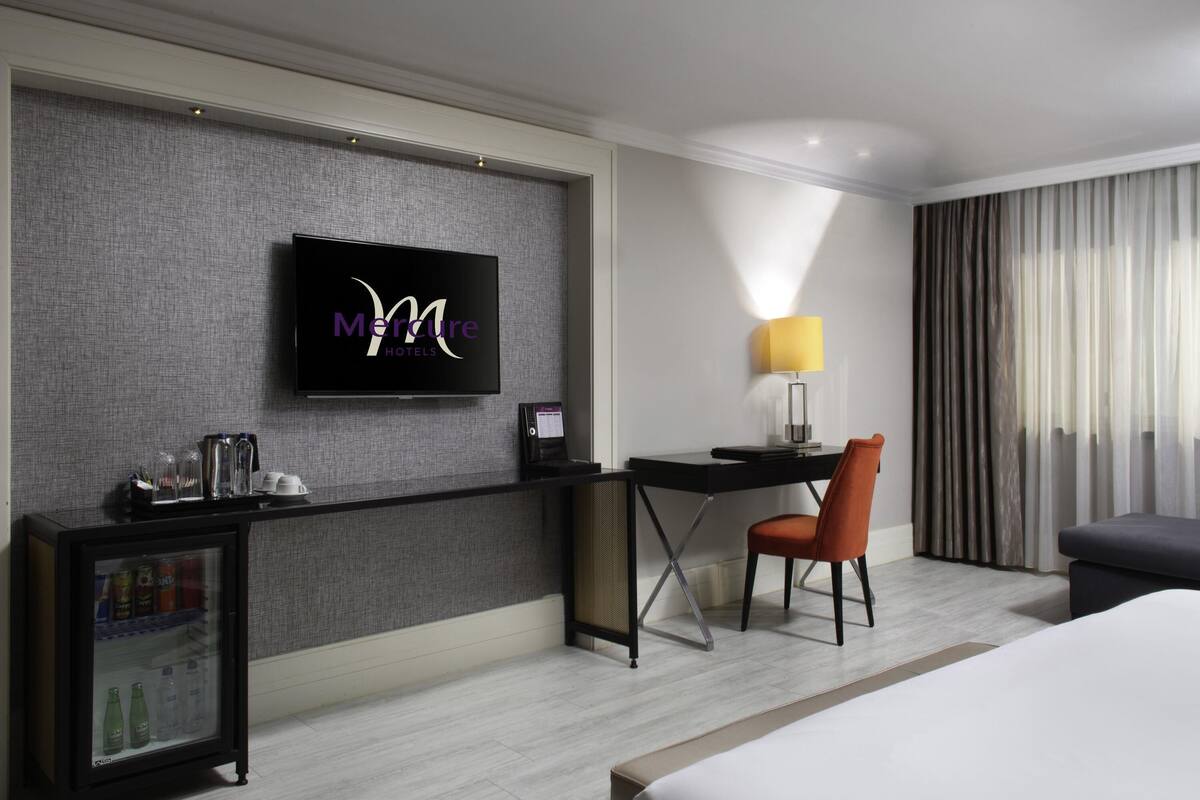 A hotel room with a bed, a desk with a chair, a television on the wall, and a minibar.
