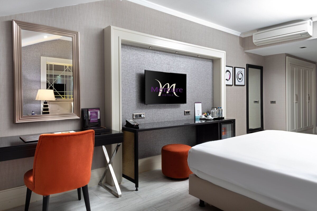 A hotel room with a bed, a desk, a TV, a mirror, and a small orange ottoman.