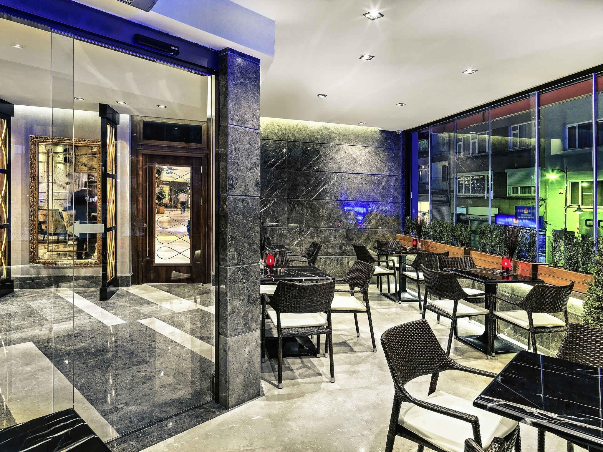 A modern hotel lobby with a glass entrance, marble flooring, and outdoor seating area with wicker furniture.
