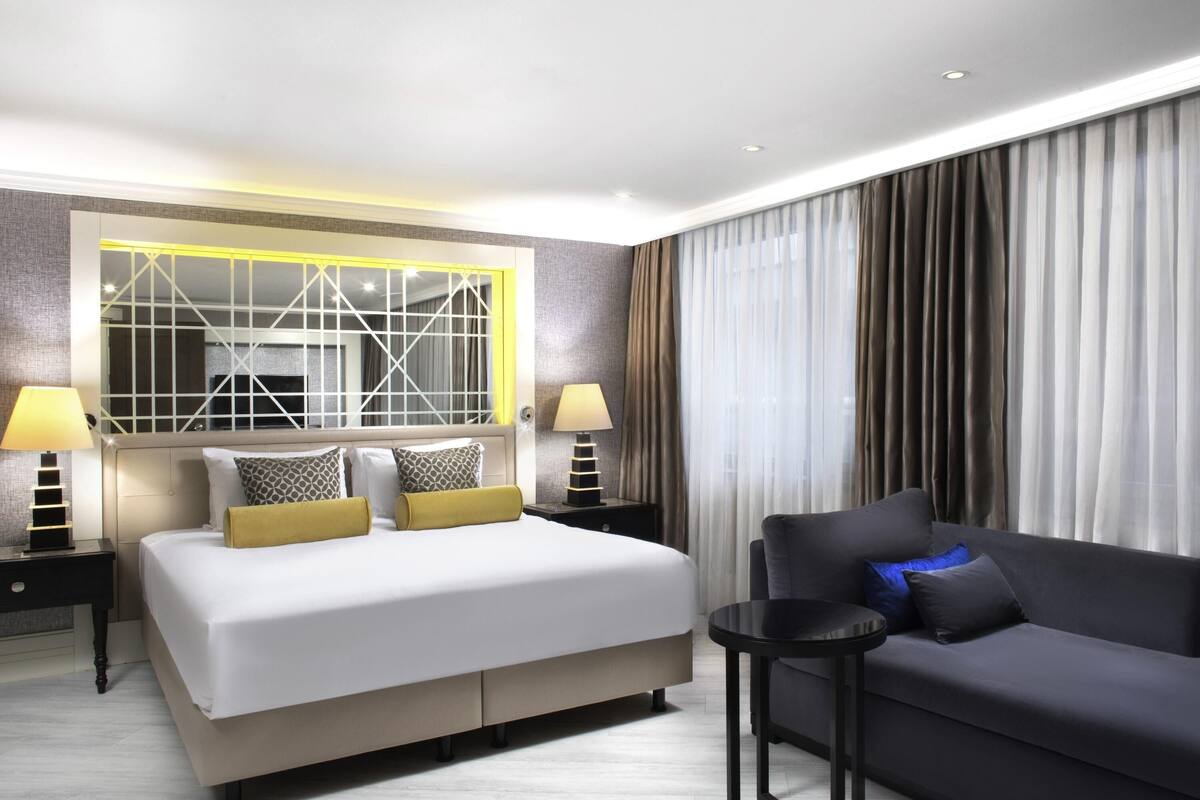 A modern hotel room with a large bed, a sofa, a small table, and a large mirror with a geometric frame.