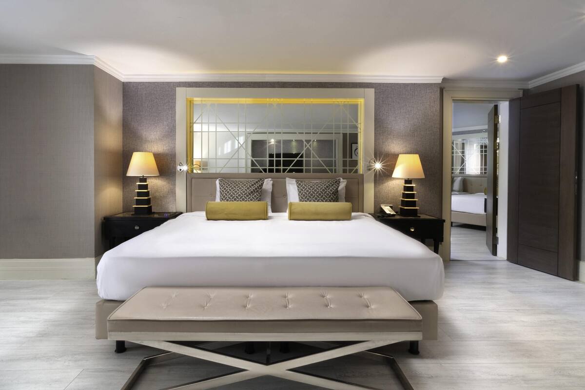 A modern hotel room with a large bed, two bedside tables with lamps, a large mirror, and a view into another room.