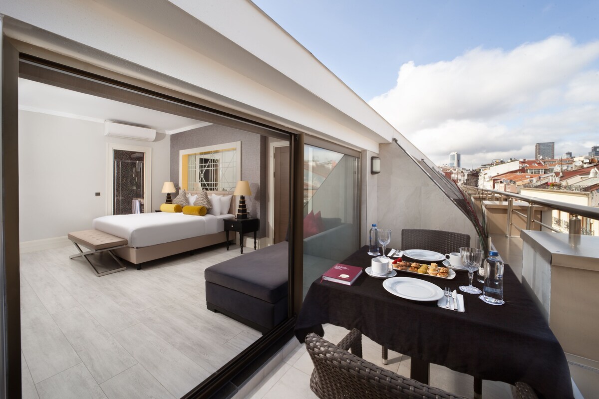 A modern hotel room with a balcony, a dining table set with plates and glasses, and a bed with a nightstand.