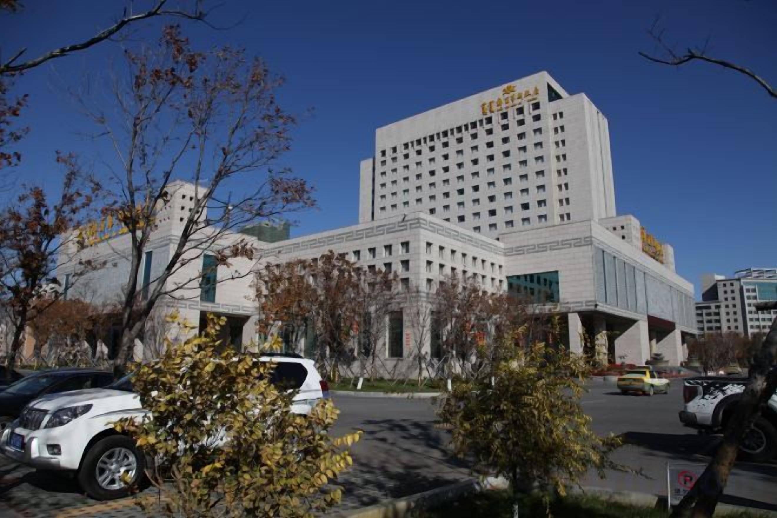 A multi-story hotel with a prominent entrance, a parking lot with several cars, and a clear blue sky.
