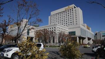 A multi-story hotel with a prominent entrance, a parking lot with several cars, and a clear blue sky.