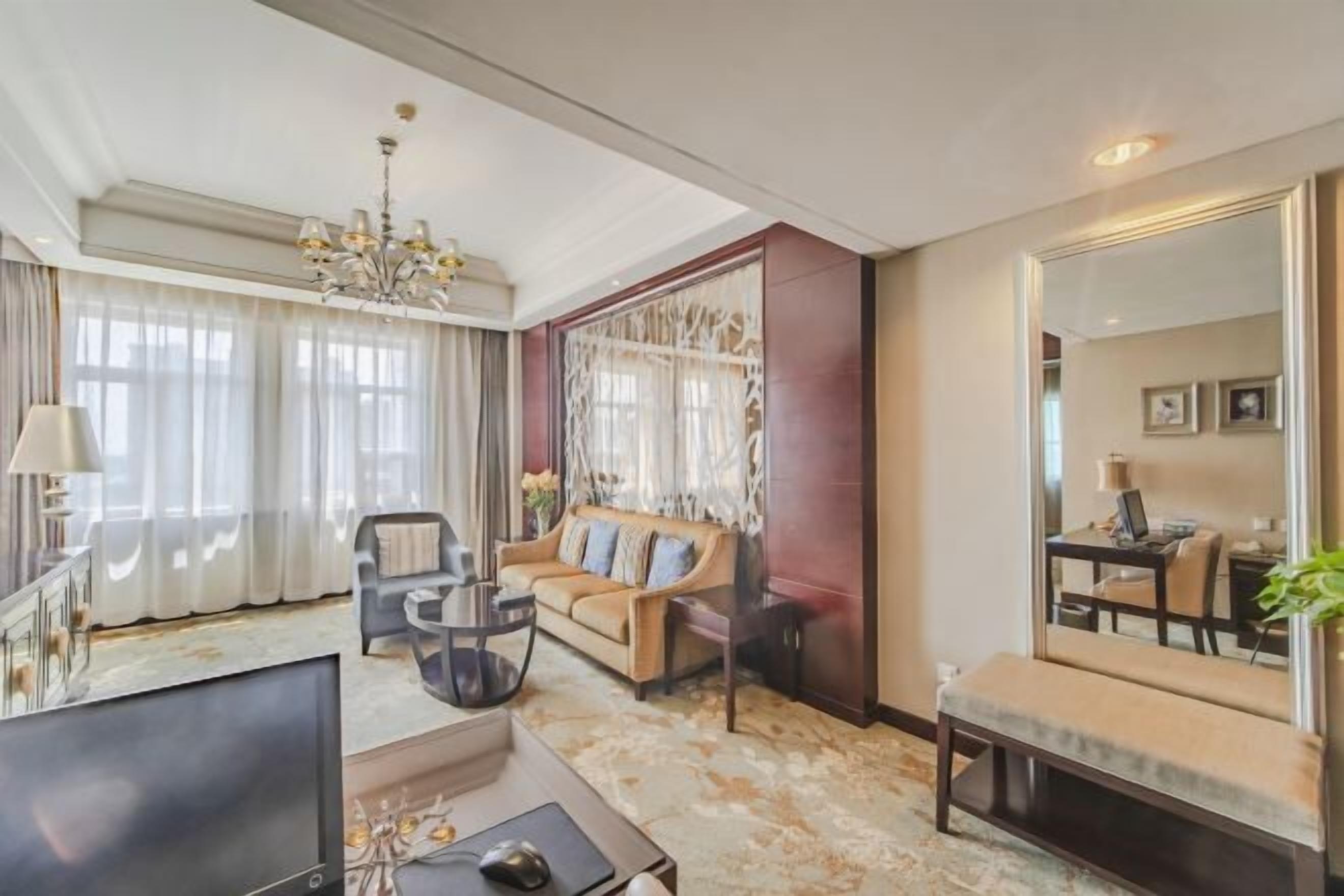 A spacious living room with a large sofa, a coffee table, and a TV. There is a chandelier, a large mirror, and a window with curtains.