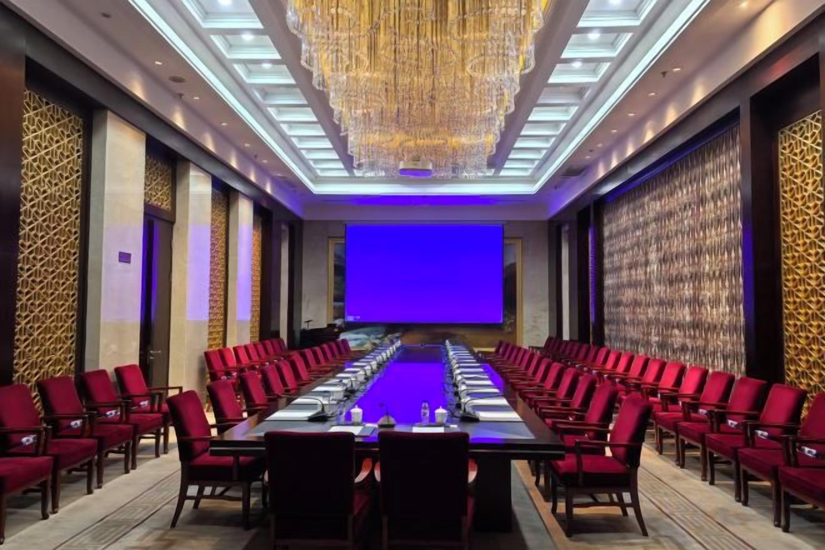 A conference room with a long table, red chairs, and a large screen.