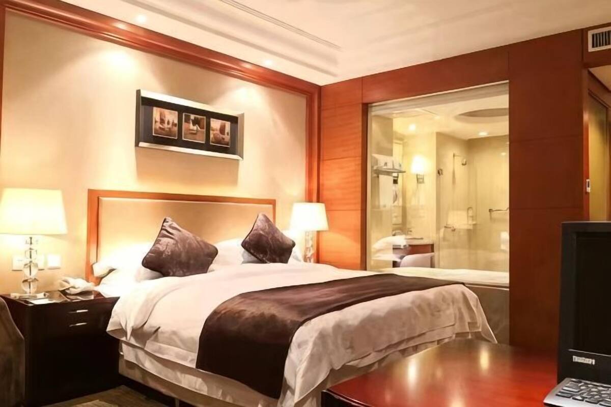 A hotel room with a bed, bedside lamps, a television, and a bathroom visible through a glass door.
