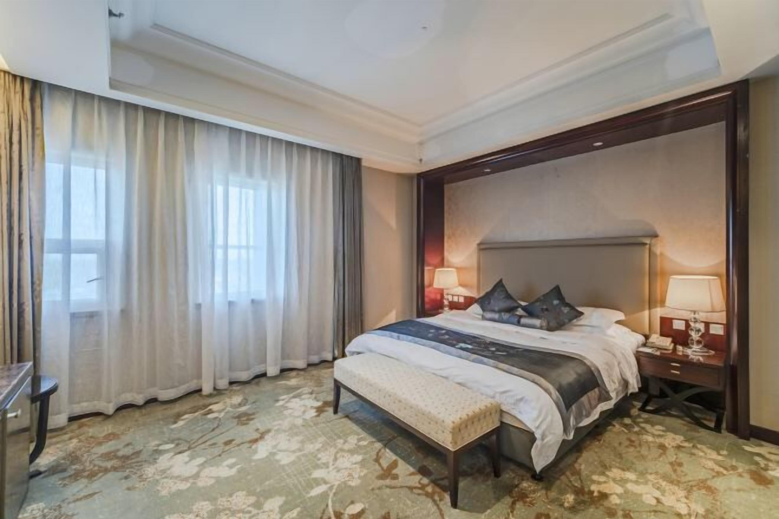 A hotel room with a large bed, two bedside tables with lamps, a bench, and a large window with curtains.