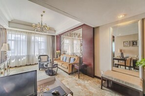 A spacious living room with a large sofa, a coffee table, and a TV. There is a chandelier, a large mirror, and a window with curtains.