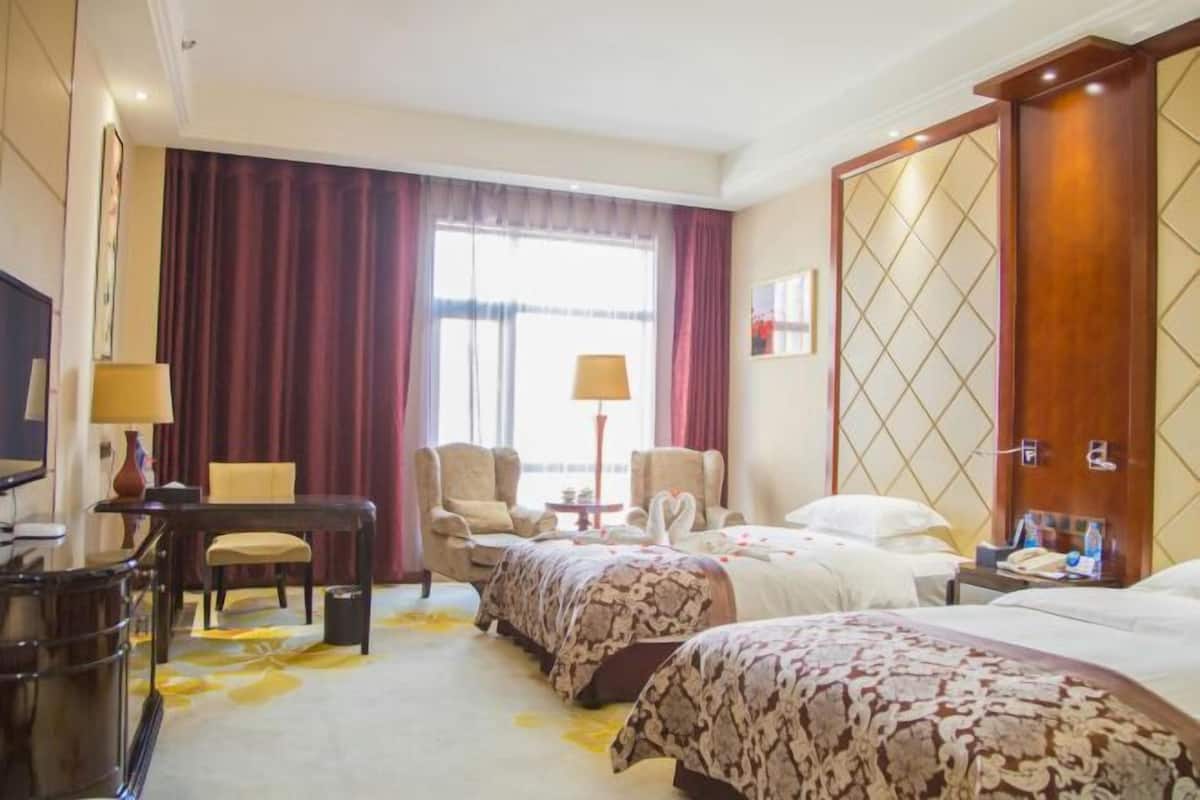 A hotel room with two beds, a desk, a chair, a TV, and a large window with curtains.