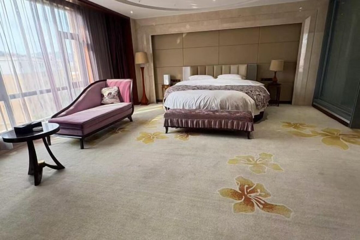 A hotel room with a large bed, a sofa, a small table, and a large window with curtains.