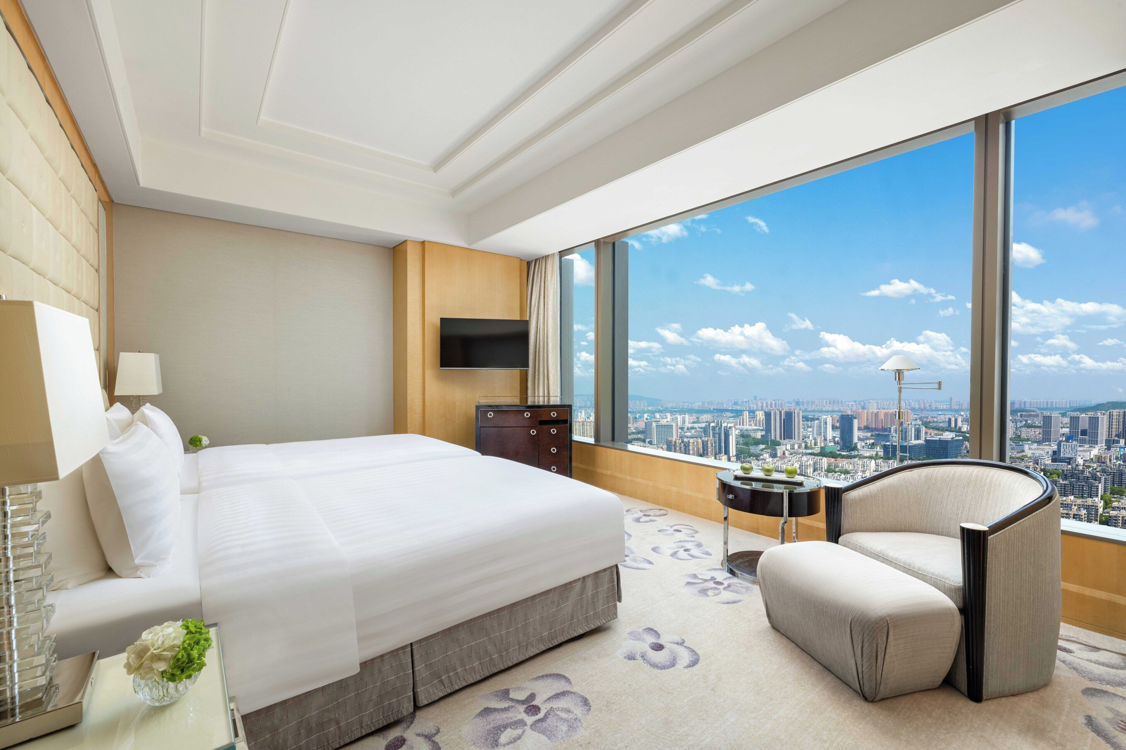 A modern hotel room with a large bed, a seating area, a flat-screen TV, and a city view through large windows.