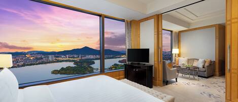 A hotel room with a large window offering a cityscape view, a bed, a TV, a desk, and a seating area.