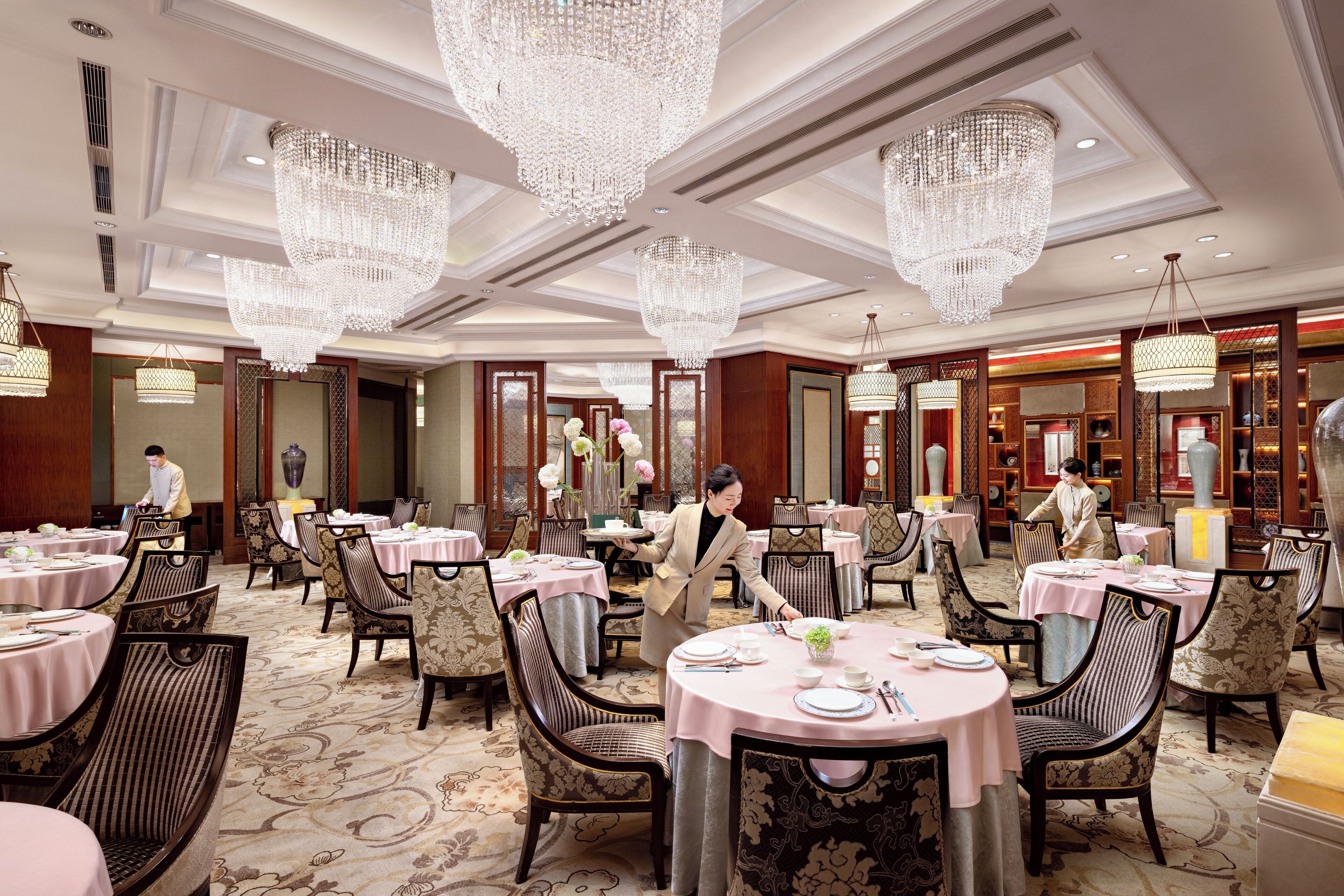 A spacious hotel dining area with round tables, elegant chandeliers, and classic decor.