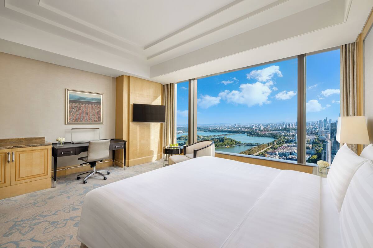 Ein Hotelzimmer mit einem großen Bett, einem Schreibtisch, einem Sessel, einem Fernseher und Blick auf eine Stadtlandschaft durch große Fenster.