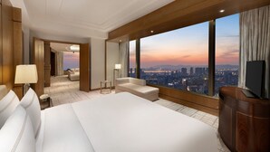 Habitación de hotel con una cama grande, un sofá, televisión y vista a la ciudad al atardecer.